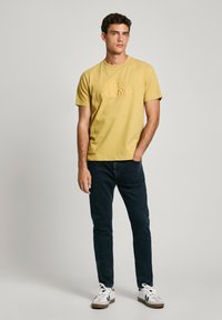 Yellow cotton t-shirt with embossed logo, paired with dark blue jeans and white sneakers. Model standing with arms relaxed at sides.