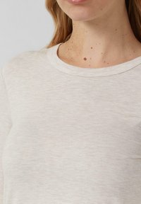 Zbliżenie na osobę w jasnobeżowej koszulce z okrągłym dekoltem, ukazujące szyję i częściowo twarz z jasnymi piegami i znamionami.