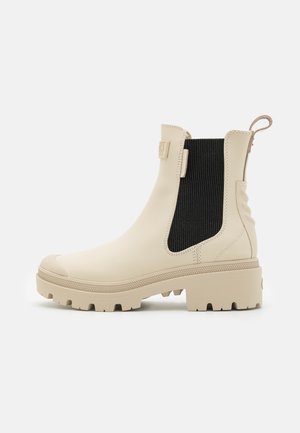 Beige waterproof Chelsea boots with black elastic side panels, rubber sole, and a textured back tab for easy wear. Chunky design with grip.