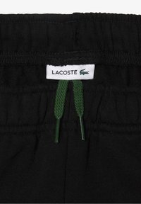 Musta joustava vyötärönauha, jossa on vihreät narut; varustettu Lacoste-logolla. Materiaali näyttää olevan pehmeää ja hengittävää. Ei lisäkuvioita näkyvissä.