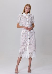 Robe en dentelle blanche avec des manches courtes, un col, une fermeture à boutons à l'avant et des motifs floraux. Dispose d'une taille ceinturée et de deux poches.