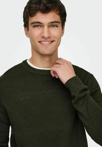 Suéter texturizado en verde oscuro con cuello redondo, que presenta un patrón de panal y puños acanalados. La tela parece suave y acogedora.