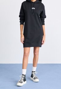 Svart hoodieklänning med ribbade mudd och nederkant, med en liten vit logotyp på bröstet. Matchas med svarta mönstrade högsneakers.