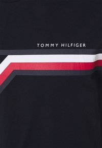 Námořnická modrá tričko s horizontálním grafickým designem s červenými, bílými a šedými pruhy. Nad nimi je tištěno jméno značky "TOMMY HILFIGER".