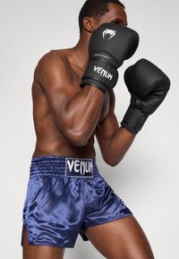 Fekete boxkesztyűk "Venum" logóval, amelyet egy személy visel, aki tengerészkék szatén boxnadrágot visel, mely elasztikus derékpánttal és oldalsó hasítékokkal rendelkezik.
