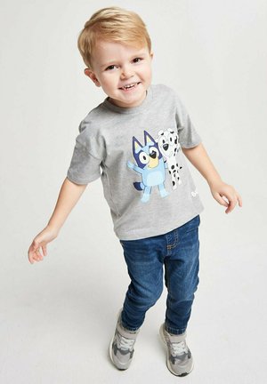 Smiling young boy with blonde hair wearing a grey cartoon character t-shirt, blue jeans, and grey sneakers, standing against a plain background.