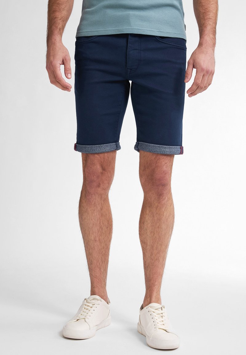 Pantalones cortos de mezclilla azul marino con dobladillo enrollado, que presentan un forro gris en contraste. Combinados con zapatillas blancas sobre un fondo neutro.