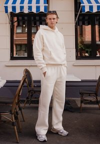 Kremfarget hettegenser med frontlomme og matchende sweatpants. Mykt stoff, avslappet passform, og parret med grå joggesko.