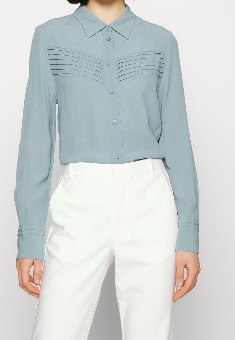 Chemise boutonnière bleu clair avec col, présentant des détails de plis horizontaux sur la poitrine, associée à un pantalon blanc taille haute.