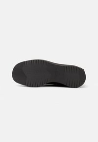 Suela de zapato negra con un patrón de goma texturizada, que presenta una disposición en zigzag y ranuras horizontales para mejorar la tracción. Logo de la marca impreso cerca del talón.