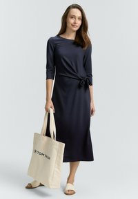 Mörkblå midi-klänning med trekvartsärmar och sidoknytning. Modell som håller en beige tote-väska med "TOM TAILOR"-logotyp. Platta sandaler.