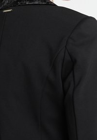 Blazer noir avec un col texturé et des épaules structurées, composé d'un tissu lisse, des coutures discrètes, et un petit logo argenté à l'arrière.