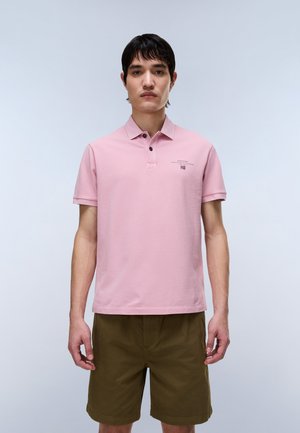 Joven de pie vistiendo una camisa polo de manga corta de color rosa claro con logo y pantalones cortos verde oliva sobre un fondo claro y liso.