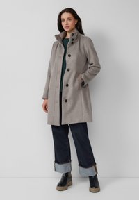 Lichtgrijze wollen jas met een hoge kraag, zes donkere knopen en zijzakken. Draagt een groene trui eronder en is gecombineerd met donkere jeans en laarzen.