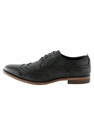 Chaussure brogue en cuir noir avec des détails perforés, bout pointu et accent de semelle brune. Design à lacets et panneaux texturés sur le dessus.