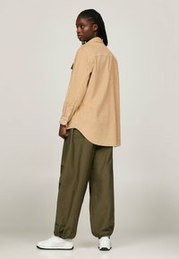 Camisa de botonadura de pana en color marrón, con un dobladillo curvado, combinada con pantalones cargo holgados en color verde oliva y zapatillas blancas, vista desde la parte posterior.