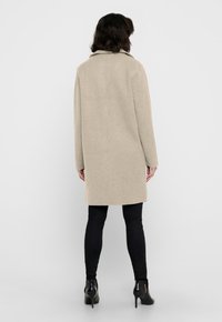 Cappotto beige in lana con taglio dritto, maniche lunghe e colletto a scollatura. La parte posteriore presenta un design pulito e si ferma poco sopra il ginocchio. Pantaloni neri e tacchi completano il look.