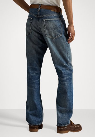 Man wearing faded blue straight-leg jeans and brown moccasin shoes, standing against a plain white background.