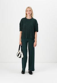 Zeleni plisirani top sa širokim rukavima, uparen s odgovarajućim hlačama. Model drži malu bijelu i crnu torbicu. Jednostavne crne cipele.