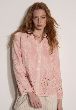 Red-haired woman wearing a pink embroidered blouse with floral patterns and scalloped edges, standing against a plain light background.