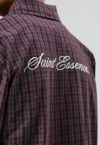 Chemise à carreaux bordeaux avec des accents bleus et blancs. Présente un texte brodé "Saint Essence" à l'arrière, près du col. Matière en coton.