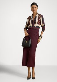 Lang burgunderfarget skjørt laget av teksturert stoff, kombinert med en mønstret bluse i mørke toner. En svart håndveske holdes, komplettert med høye hæler.