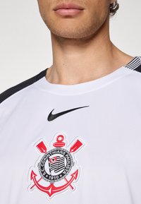 Hvitt skjort med svarte detaljer, som har Nike-logoen og et detaljert rødt og svart klubbvåpen med en anker og kryssede padler.