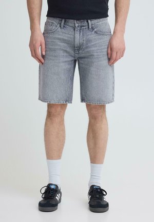 Mann iført lysegrå denimshorts, svarte joggesko med hvite striper og hvite sokker, står mot ensfarget bakgrunn.