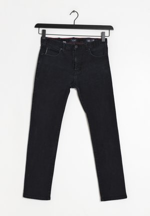 Jean noir slim-fit suspendu sur un cintre noir contre un fond blanc, avec poches avant et fermeture à boutons.