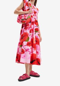 Květinové midi šaty v růžové barvě s červenými a zelenými vzory, s volánkovým designem. Oblečené s odpovídající kabelkou a růžovými plošinovými sandály.