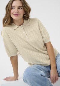 Beige polo svetr s texturovaným diamantovým vzorem, krátkými rukávy a třemi knoflíky na límečku. V kombinaci s světle modrými džíny.