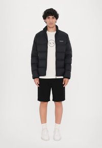 Sort pufferjakke med horisontale kviltede sektioner, lynlåslukning og stående krave, båret over en hvid grafisk t-shirt og sorte shorts.