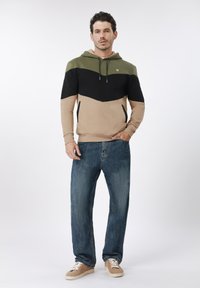 Φούτερ με κουκούλα που διαθέτει χρωματικά μπλοκ σε πράσινο, μαύρο και μπεζ· απαλό ύφασμα με άνετη γραμμή. Συνδυάζεται με χαλαρά μπλε τζιν και ταμπά αθλητικά παπούτσια.