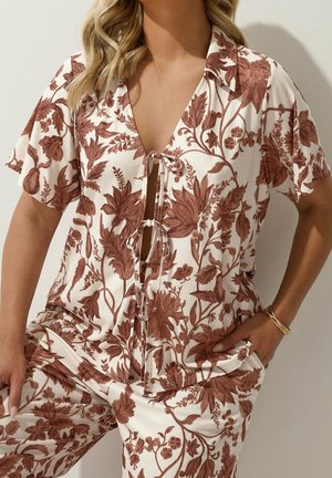 Femme portant un ensemble de vêtements d'intérieur crème avec imprimé floral marron, manches courtes, col et détail noué à l'avant, posant avec une main dans la poche.