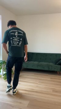 Tumši zaļš T-krekls ar baltu grafiku, melni kokvilnas bikses un sporta apavi. Olīvzaļš tuftēts dīvāns labi apgaismotā telpā ar koka grīdu.