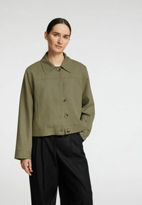 Chaqueta recortada de color verde oliva con cuello puntiagudo, cierre de cinco botones y dos bolsillos laterales. Llevada sobre pantalones negros. Textura de tela suave.