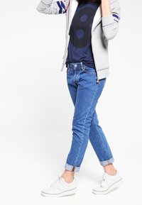 Felpa grigia chiara con zip e strisce blu, t-shirt navy con grande stampa circolare e jeans blu con risvolto, abbinati a sneaker bianche.