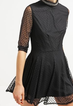 Zwarte jurk met doorschijnende mouwen en rok-overlay met polka dots, hoge halslijn met kralen, getailleerde taille en uitlopende rok.