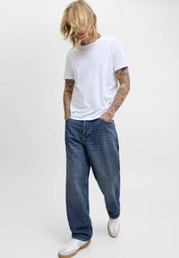 T-shirt en coton blanc, coupe décontractée, assortie à un jean bleu denim ample. Chaussures blanches avec des semelles marron. Tatouages visibles sur les bras.