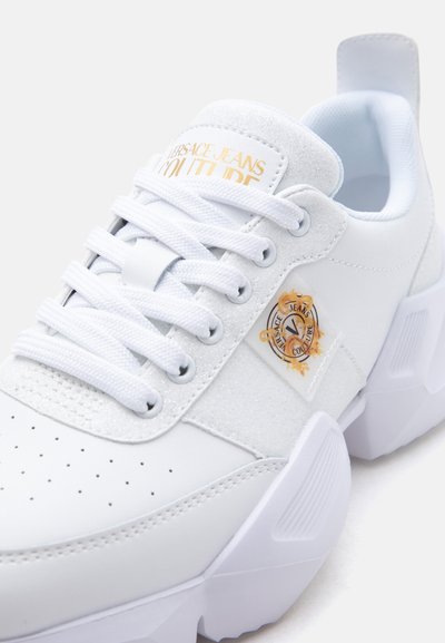 Baskets blanches épaisses avec lacets blancs, embout perforé et logo Versace Jeans Couture orné d'accent doré sur le côté et la languette.