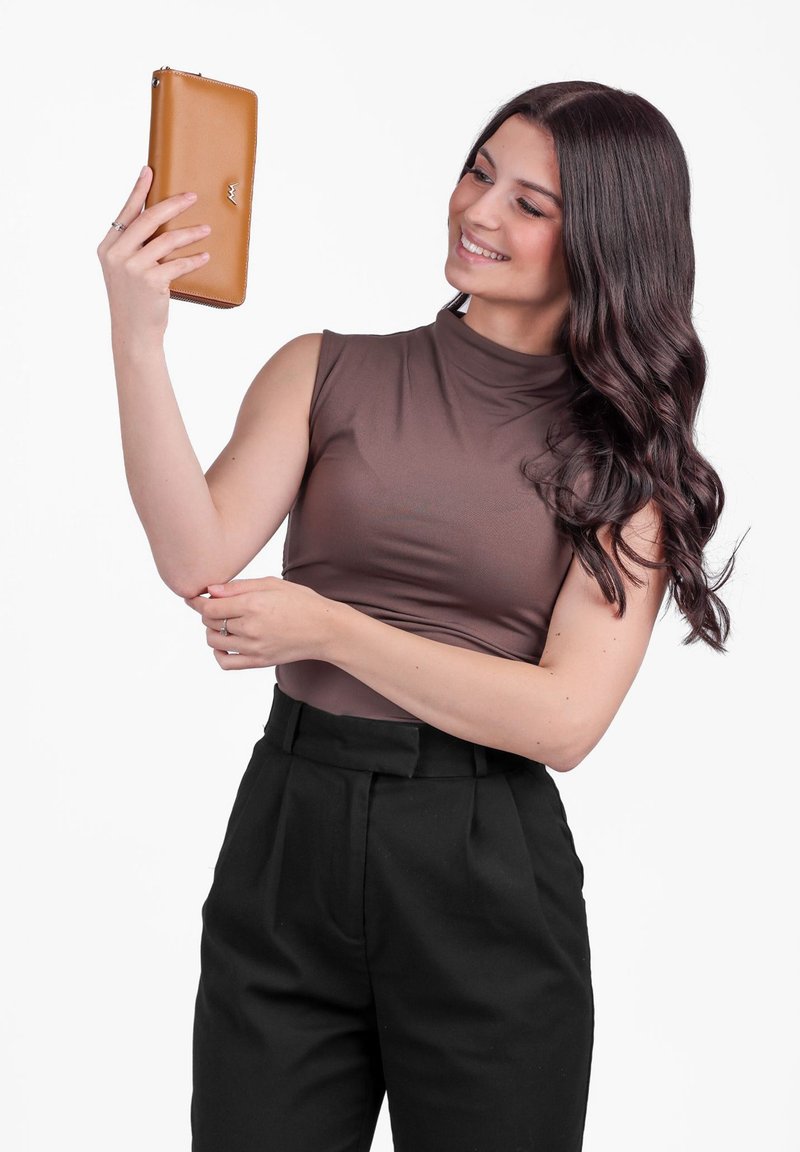 Brown zippered wallet held by a person wearing a sleeveless, high-neck brown top and black tailored pants. Background is plain white.