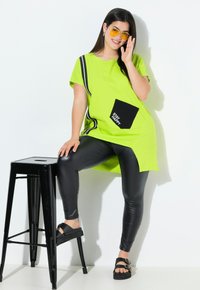 Túnica de gran tamaño en verde lima con bolsillo negro, texto gráfico y raya lateral, combinada con leggins negros y sandalias gruesas.
