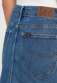 Zila džinsu svārki ar augstu jostasvietu, ar zelta šuvēm, brūnu ādas etiķeti aizmugurē un mazu etiķeti kabatā.