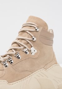 Bota ankle de ante beige con un upper texturizado, herrajes plateados y suela de goma color crema. Los cordones y ojales proporcionan un ajuste seguro.