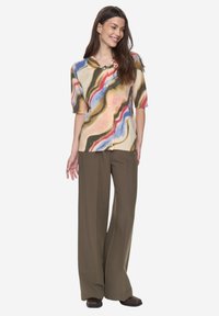 Bunte gemusterte Kurzarmpullover-Strickjacke mit Knöpfen, die Spiralen in Pink, Blau und Gelb zeigt. Kombiniert mit lockeren olivgrünen Hosen.