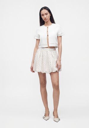 Jeune femme aux longs cheveux foncés portant un top court blanc à manches courtes avec des attaches dorées et une mini-jupe plissée blanche à motifs.