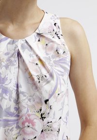 Ärmeles blus i lätt tyg med blommönster i pastellfärger och svarta accenter; plisserad halsringning ger textur och detalj.