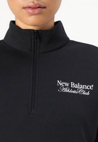 Zwarte atletische trui met een kwart ritssluiting. Heeft een hoge halslijn en een wit "New Balance Athletic Club" logo op de borst. Soepele textuur.