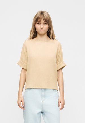 Ung kvinde med glat lysebrunt hår iført en beige top med korte ærmer og lyseblå højtaljede jeans, stående foran en ensfarvet baggrund.