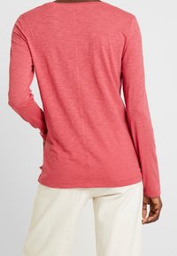 Långärmad topp i melerad korallfärgad tyg. Har en rund halsringning, sömdetaljer och en rundad nederkant. Bärs med ljusa byxor.